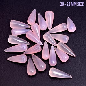 Può includere: Una collezione di gemme a forma di goccia, rosa pallido, ciascuna di circa 20-22 mm, su una superficie scura. Le pietre traslucide hanno un aspetto lucido e liscio, con una sottile sfumatura di colore.