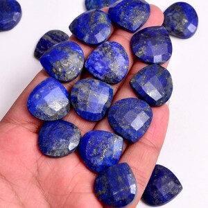 Puede incluir: Colección de piedras preciosas de lapislázuli azul profundo facetadas. Las piedras tienen forma de corazón y presentan motas doradas. Algunas piedras se sostienen en una mano, otras están sobre una superficie blanca.