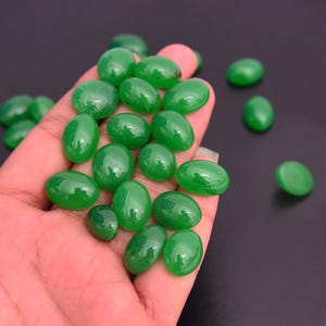 Op de afbeelding: Een handvol gepolijste, ovaalvormige groene edelstenen. De stenen variëren enigszins in grootte en hebben een glanzend, reflecterend oppervlak. De edelstenen hebben een levendige, rijke groene kleur en voelen glad aan. De achtergrond is donker en neutraal.