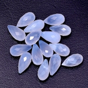 Puede incluir: Un conjunto de gemas en forma de l&aacute;grima de color azul p&aacute;lido, con superficies facetadas. Las gemas est&aacute;n dispersas sobre un fondo oscuro y texturizado, mostrando su calidad transl&uacute;cida y sutiles variaciones de color. Ideales para la fabricaci&oacute;n de joyas.