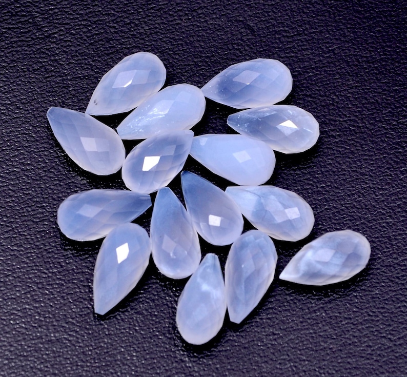 Puede incluir: Un conjunto de gemas en forma de l&aacute;grima de color azul p&aacute;lido, con superficies facetadas. Las gemas est&aacute;n dispersas sobre un fondo oscuro y texturizado, mostrando su calidad transl&uacute;cida y sutiles variaciones de color. Ideales para la fabricaci&oacute;n de joyas.