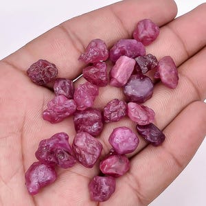May include: A collection of raw, uncut ruby gemstones in varying shades of pink and red. The gemstones have a rough, natural texture and irregular shapes. The gems are scattered across a surface, showcasing their natural beauty.