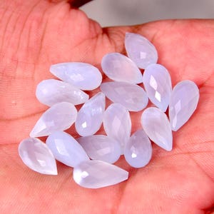 Puede incluir: Un conjunto de gemas en forma de l&aacute;grima, de color azul claro, con superficies facetadas. Las gemas se muestran sobre un fondo color piel, resaltando su cualidad transl&uacute;cida y sutiles variaciones de color. Adecuado para la creaci&oacute;n de joyas.