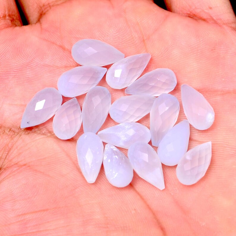 Puede incluir: Una colecci&oacute;n de gemas en forma de l&aacute;grima de color lavanda p&aacute;lido con superficies facetadas. Las gemas est&aacute;n dispersas sobre una superficie rosa claro, mostrando su calidad transl&uacute;cida y sutiles variaciones de color. Las gemas son de varios tama&ntilde;os.