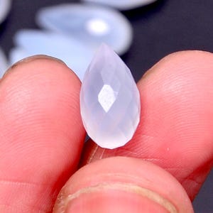 Puede incluir: Una gema facetada en forma de l&aacute;grima, de color p&aacute;lido y transl&uacute;cido. La gema se sostiene entre los dedos, mostrando sus facetas pulidas y sutiles variaciones de color. La forma de la gema es alargada, con una punta.