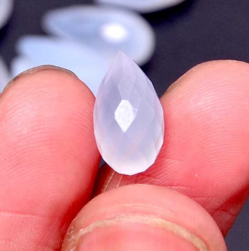 Puede incluir: Una gema facetada en forma de l&aacute;grima, de color p&aacute;lido y transl&uacute;cido. La gema se sostiene entre los dedos, mostrando sus facetas pulidas y sutiles variaciones de color. La forma de la gema es alargada, con una punta.