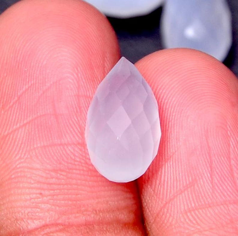 Puede incluir: Una gema facetada en forma de l&aacute;grima, de color lavanda p&aacute;lido. La gema se sostiene entre los dedos, mostrando su intrincado corte y cualidad transl&uacute;cida. Las facetas capturan la luz, creando un efecto brillante. El tema general es joyer&iacute;a y piedras preciosas.