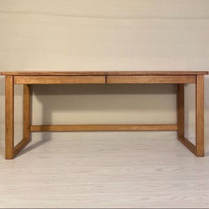 May include: A wooden desk with a natural finish. The desk has a rectangular top with two drawers and a simple, sturdy frame. The desk is made of light-colored wood and has a minimalist design, suitable for a home office or workspace.