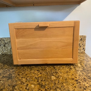 May include: A wooden bread box with a hinged door and a small handle. The box is sitting on a granite countertop.