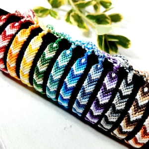 May include: A set of 10 colourful friendship bracelets with chevron patterns. The bracelets are made with white and various colours of embroidery floss. The bracelets are arranged in a rainbow order, starting with red and ending with orange.