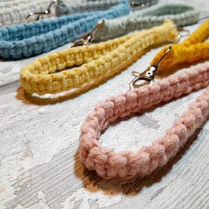 May include: Close-up of several macrame lanyards in various colours, including yellow, pink, blue, and grey. Each lanyard features a woven design and a silver-coloured clasp for attaching keys or an ID badge.