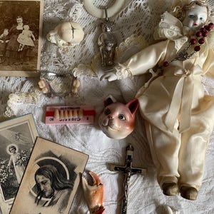 May include: A collection of vintage items including a porcelain doll in cream clothing, a silver bear rattle, a small skull, a set of dentures, a cat head figurine, a crucifix, a crab claw, an animal jawbone, and two sepia-toned religious cards.