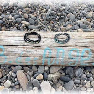 Könnte beinhalten: Zwei schwarze Perlenarmbänder mit goldenen und türkisfarbenen Akzenten auf einem verwitterten Holzbrett mit den Worten "Beach Vibes" in Blau.