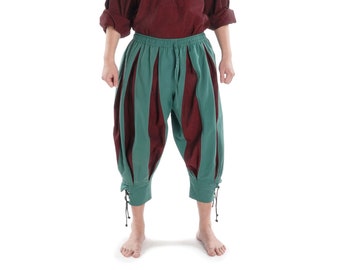 May include: A man wearing a burgundy shirt with a lace-up neckline and green and burgundy pants with a unique design. He is wearing a burgundy hat.