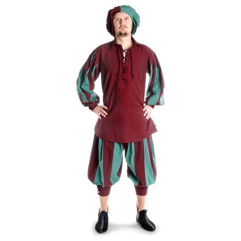 May include: A man wearing a red and green medieval costume with a green hat. He is standing with his hands on his hips.