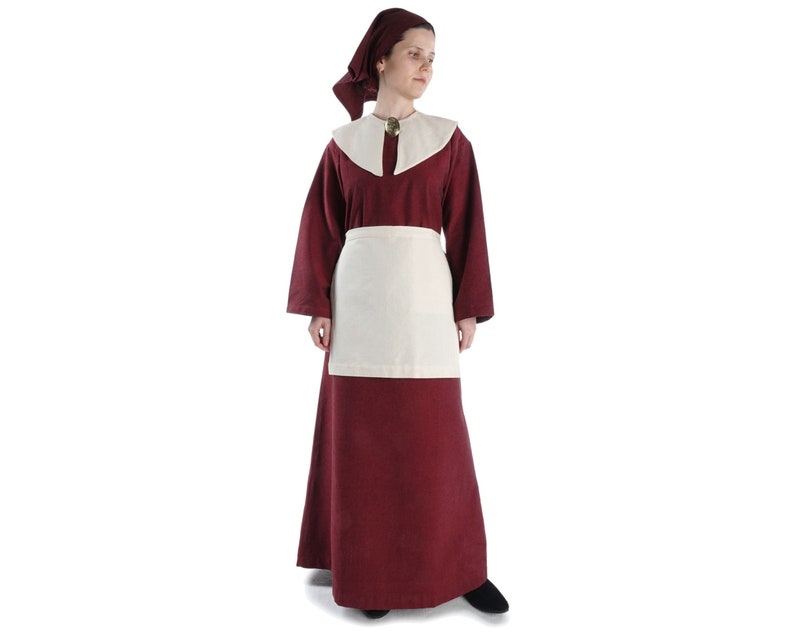 May include: A woman wearing a long, burgundy dress with a white apron and a burgundy head covering. She is standing on a white background.
