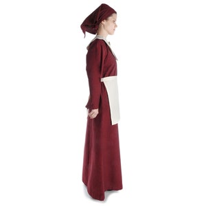 May include: A woman wearing a long, burgundy dress with a white apron and a burgundy headscarf.