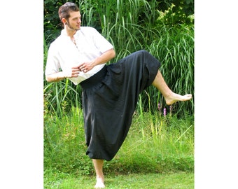 May include: A person wearing a white shirt with a lace-up neckline and black, loose-fitting pants. The person is standing in a grassy area with tall green plants.