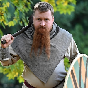 Könnte beinhalten: Eine Person in historischer Kleidung, die eine Axt über der Schulter und einen Schild hält. Die Person hat einen langen, roten Bart und trägt einen grau-schwarz gemusterten Schal. Der Schild hat ein kreisförmiges Design mit orangefarbenen, grünen und braunen Segmenten.