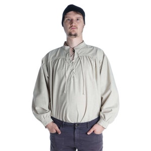 Pode incluir: Camisa bege de manga comprida com decote com atacadores e gola mandarim. A camisa tem detalhes franzidos nos ombros e punhos. A pessoa na imagem est&aacute; usando um gorro preto e cal&ccedil;as azul-escuras.