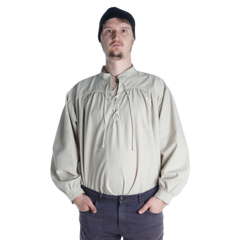 Pode incluir: Camisa bege de manga comprida com decote com atacadores e gola mandarim. A camisa tem detalhes franzidos nos ombros e punhos. A pessoa na imagem est&aacute; usando um gorro preto e cal&ccedil;as azul-escuras.