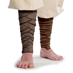 Könnte beinhalten: Braune Beinwickel mit strukturiertem Muster, getragen mit einem cremefarbenen Kleidungsstück. Die Wickel sind eng um die Unterschenkel gewickelt und erzeugen einen geschichteten Effekt. Das Bild ist vor weißem Hintergrund.