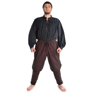 May include: A man wearing a black long-sleeved shirt with a lace-up neckline and brown pants with a drawstring waist.