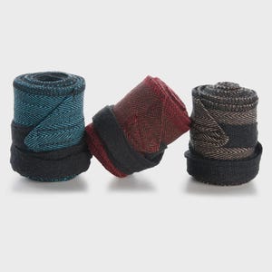 May include: Three rolled fabric wraps featuring a herringbone pattern. The first wrap is teal and black, the second is deep red and black, and the third is tan and black. Each roll is secured with a solid black fabric strap at the base.