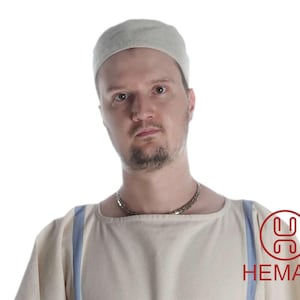 Könnte beinhalten: Ein Mann trägt eine weiße Leinen-Tunika mit blauen Trägern, eine weiße Filzmütze und eine silberne Kettenkette. Der Mann schaut direkt in die Kamera. Das Bild enthält den Text "HEMAD" in Rot.