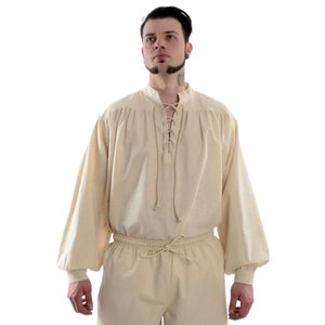 Pode incluir: Uma camisa creme em estilo renascentista com mangas compridas e volumosas e decote com atacadores. A camisa &eacute; combinada com cal&ccedil;as a condizer, ambas feitas de um tecido macio e leve. O conjunto tem cintura e punhos com cord&atilde;o.