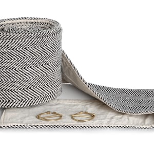 May include: A rolled-up black and white herringbone fabric with a matching extended strip. Two gold-colored brooches rest on the fabric. The fabric has a neutral-colored lining.