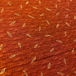 May include: Rust-colored fabric with a repeating pattern of gold-colored leaf shapes. The fabric has a textured appearance with small, dark brown dots scattered across the surface. The leaves are stylized and evenly spaced.