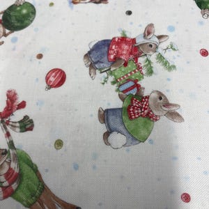 May include: White fabric with a repeating Christmas-themed pattern. The design features cartoon rabbits and reindeer wearing festive outfits and carrying gifts. Scattered snowflakes and ornaments complete the holiday scene.