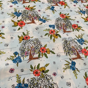 May include: Light blue fabric with a repeating pattern of stylized trees, flowers, and butterflies. The trees have brown trunks and green leaves, with clusters of red, blue, and yellow flowers. The design includes small black dots and horizontal lines.