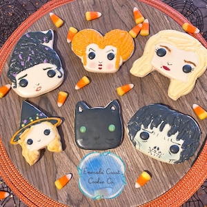 May include: A selection of Halloween-themed sugar biscuits, featuring character faces and a black cat, presented on a wooden surface with candy corn. The biscuits are intricately iced and set within an orange border.