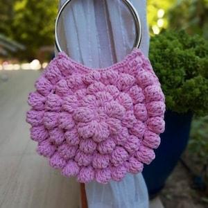 Könnte beinhalten: Eine rosa gehäkelte Handtasche mit einem silbernen Ringgriff. Die Tasche ist aus einem strukturierten, bauschigen Stoff.