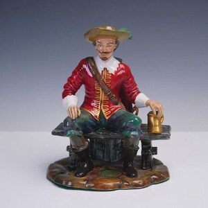 Puede incluir: Figura de cerámica de un hombre con chaqueta roja, pantalón verde y botas negras, sentado en un banco de madera. Sostiene una jarra dorada. La figura tiene un sombrero con pluma y una bandolera marrón. La base es marrón y verde.