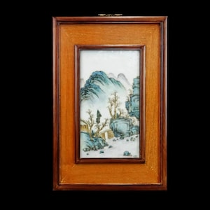 May include: A framed porcelain tile with a painted landscape scene. The tile depicts a mountain range with a small village in the foreground. The mountains are painted in shades of blue and green, while the village is painted in shades of brown and white.