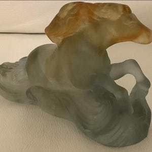 Pode incluir: Escultura em vidro fosco de um cavalo em pose reclinada. A crina e a cabeça do cavalo são âmbar, passando para um corpo translúcido com tons de cinza e azul. A escultura é detalhada e artística.