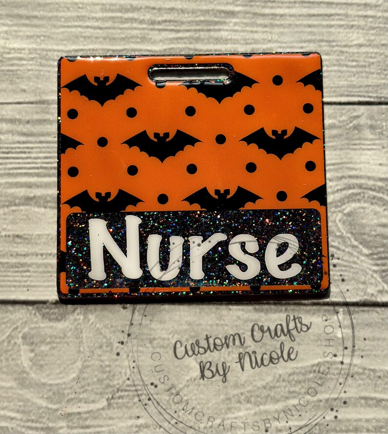 May include: Orange and black Halloween-themed badge with a black glitter background and the word "Nurse" in white letters. The badge has a hole at the top for a lanyard.