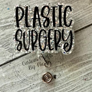 May include: A silver glitter badge holder with the text "Plastic Surgery" in black lettering. The badge holder has a clear plastic backing and a metal clip.