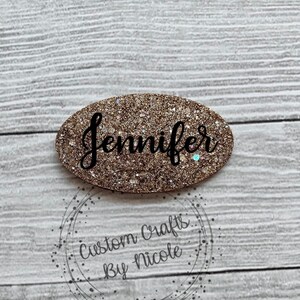 May include: Gold glitter oval with the name "Jennifer" written in black cursive lettering.