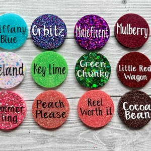 May include: Twelve round, glitter-covered, resin discs with various colours and text. The text includes names like "Tiffany Blue", "Orbitz", "Maleficent", "Mulberry", "Iceland", "Key Lime", "Green Chunky", "Little Red Wagon", "Summer Fling", "Peach Please", "Reel Worth It", and "Cocoa Bean".