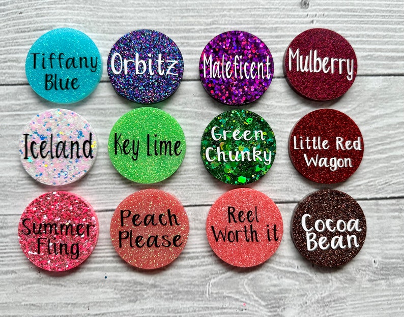 May include: Twelve round, glitter-covered, resin discs with various colours and text. The text includes names like "Tiffany Blue", "Orbitz", "Maleficent", "Mulberry", "Iceland", "Key Lime", "Green Chunky", "Little Red Wagon", "Summer Fling", "Peach Please", "Reel Worth It", and "Cocoa Bean".