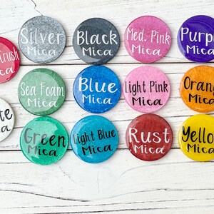 May include: A collection of 12 small round containers with different coloured mica powders. The containers are labelled with the colour and type of mica powder. The colours include: orange crush, white, sea foam, blue, light pink, orange, green, light blue, rust, yellow, silver, black, medium pink, and purple.