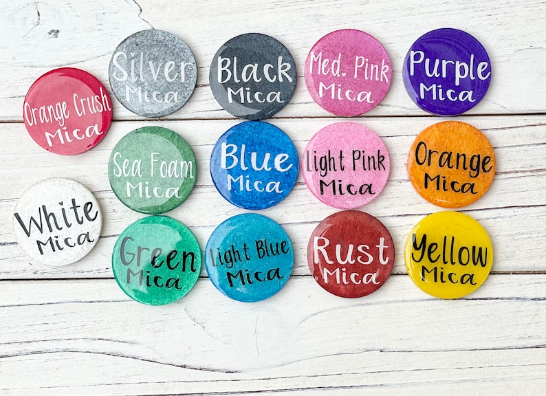 May include: A collection of 12 small round containers with different coloured mica powders. The containers are labelled with the colour and type of mica powder. The colours include: orange crush, white, sea foam, blue, light pink, orange, green, light blue, rust, yellow, silver, black, medium pink, and purple.
