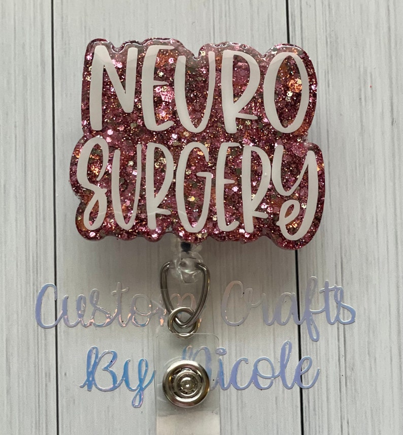 May include: Pink glitter badge holder with white "NEURO SURGERY" text. The badge holder is attached to a clear retractable badge reel.