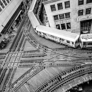 May include: A black and white aerial view of a train crossing over a network of train tracks in a city. The train is traveling over a bridge and the tracks below are in a grid pattern.
