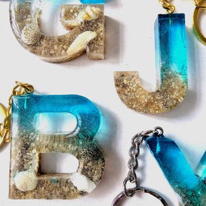 May include: Resin letter keychains featuring a gradient design that transitions from a clear blue top to a sandy bottom filled with small seashells and sand particles. Each letter includes a gold-toned metal keyring attached to the top.
