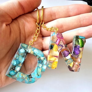 May include: A handmade keychain with the letters "D" and "N". The letters are filled with colourful stones and gold flakes, set in clear resin. The keychain has a gold chain and key ring.